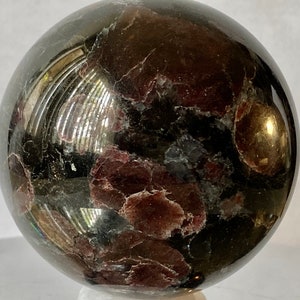 May include: A polished black sphere with red and brown inclusions. The sphere is made of a natural stone, possibly a type of garnet.