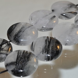 May include: A strand of round, smooth, black and white gemstone beads. The beads are translucent and have a cloudy appearance. The beads are strung on a black cord.
