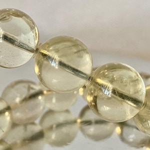 Puede incluir: Primer plano de una hebra de cuentas de piedras preciosas de color amarillo claro. Las cuentas son redondas y lisas, con un ligero brillo. Las cuentas están enhebradas en un alambre de plata.