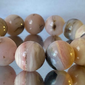 May include: A bracelet made of pink and white agate beads. The beads are round and have a smooth, polished finish. The bracelet is a light pink color with white streaks throughout.