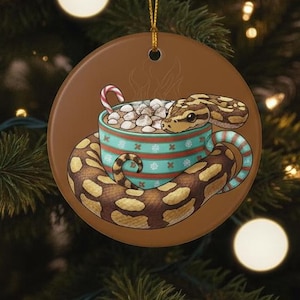 May include: A round ceramic Christmas ornament featuring a snake coiled around a teal mug filled with marshmallows and a candy cane. The ornament has a brown background and a gold string for hanging.