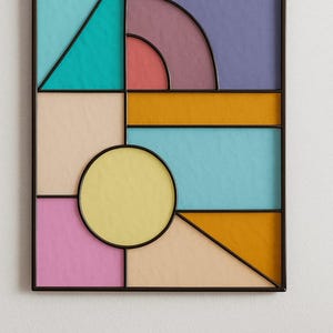 Puede incluir: Arte mural geométrico con marco negro. La obra presenta varias formas y colores, incluyendo turquesa, morado, amarillo, rosa y naranja. El diseño incluye triángulos, rectángulos y un círculo, creando una estética moderna.