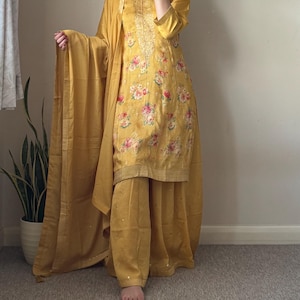May include: Mustard yellow salwar kameez outfit. The tunic has a floral print with pink and red roses. The set includes matching trousers and a dupatta. The outfit has gold embroidery around the neckline and on the tunic.