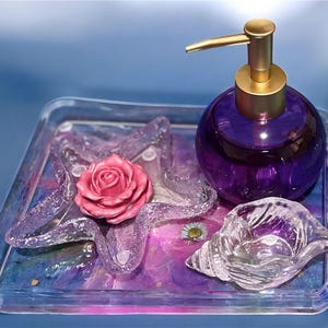 May include: A decorative tray with a purple and blue marbled design, holding a purple glass soap dispenser with a gold pump, a pink rose, and a clear seashell-shaped dish. The tray also features clear, wavy decorative elements.