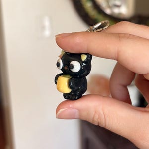 May include: A black cat-shaped keychain with large white eyes and yellow accents, holding a small yellow square. The keychain has a silver clasp. The cat is approximately 1.5 inches tall.