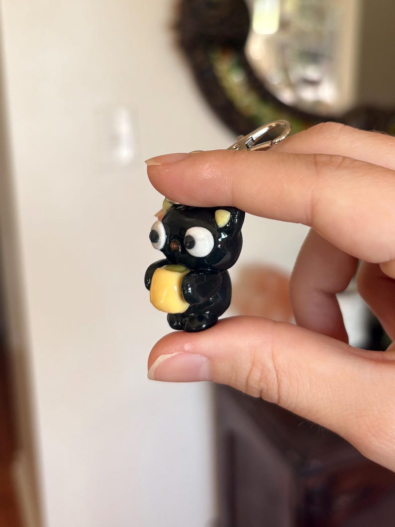 May include: A black cat-shaped keychain with large white eyes and yellow accents, holding a small yellow square. The keychain has a silver clasp. The cat is approximately 1.5 inches tall.
