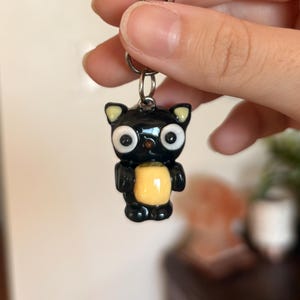 May include: Black cat keychain with large white eyes, yellow ears, and a yellow belly. The cat is holding a yellow object. The keychain has a silver clasp.