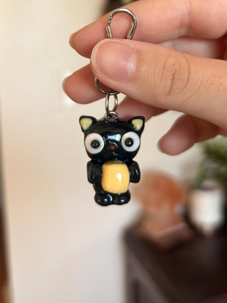 May include: Black cat keychain with large white eyes, yellow ears, and a yellow belly. The cat is holding a yellow object. The keychain has a silver clasp.