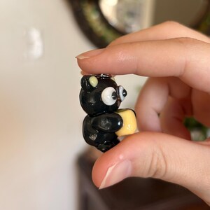 May include: A small, black bear figurine with large, white eyes and a yellow square. The bear is holding the yellow square. The figurine is attached to a small silver keychain.