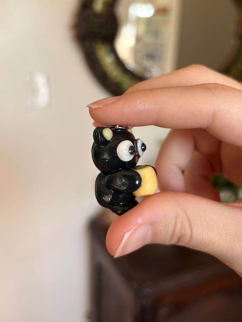 May include: A small, black bear figurine with large, white eyes and a yellow square. The bear is holding the yellow square. The figurine is attached to a small silver keychain.