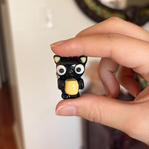 May include: A small, black cat figurine with large, white eyes and a yellow belly. The cat has small, yellow-tipped ears and is held between fingers. The figurine is likely made of a glossy material.