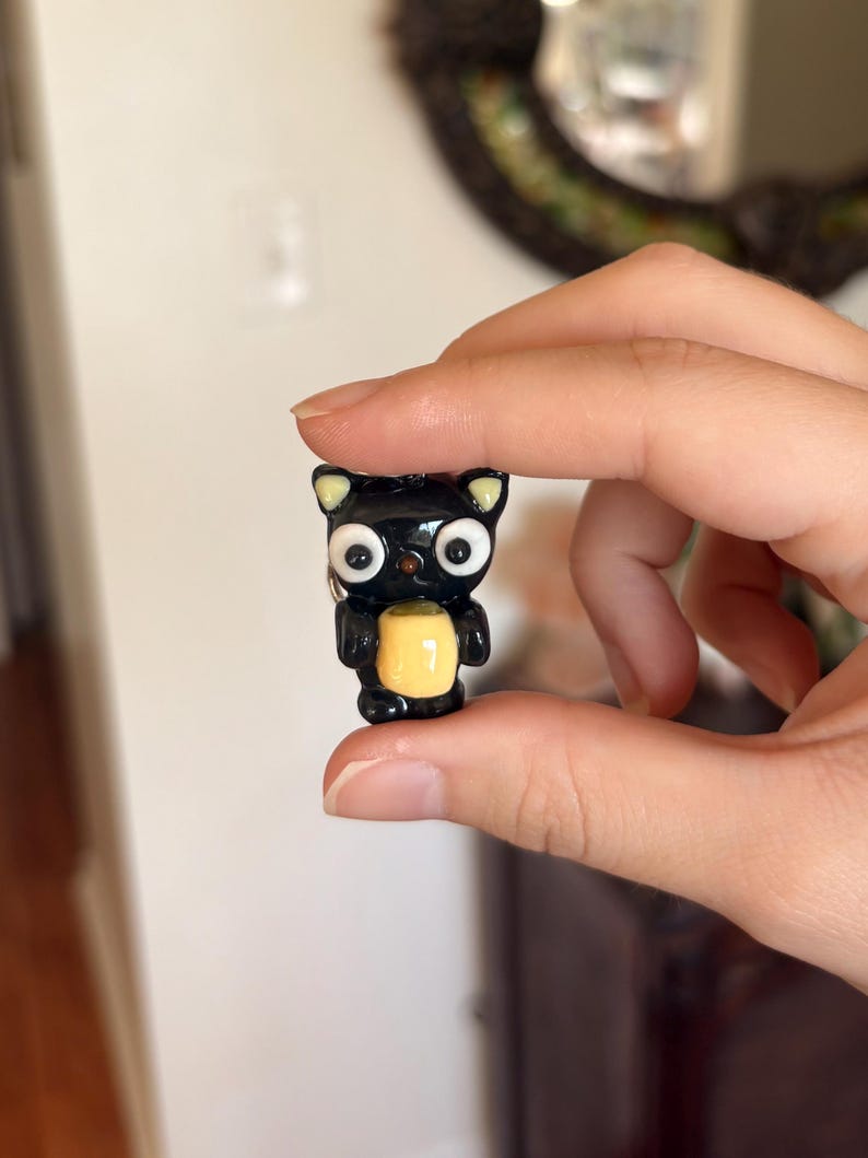 May include: A small, black cat figurine with large, white eyes and a yellow belly. The cat has small, yellow-tipped ears and is held between fingers. The figurine is likely made of a glossy material.