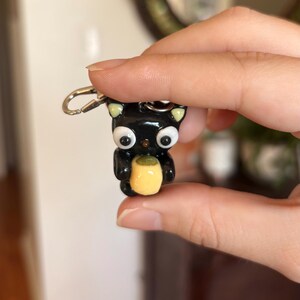 May include: A black cat keychain with large white eyes, yellow ears, and a yellow object held in its paws. The keychain is made of a glossy material and has a silver clasp.