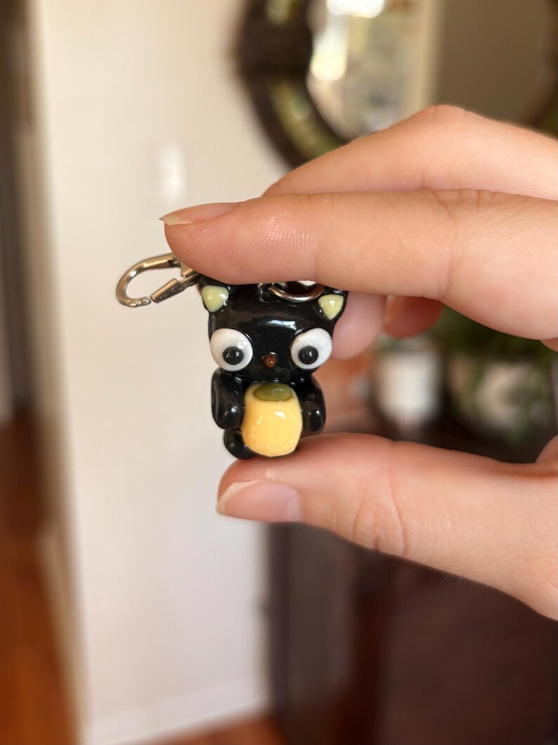 May include: A black cat keychain with large white eyes, yellow ears, and a yellow object held in its paws. The keychain is made of a glossy material and has a silver clasp.