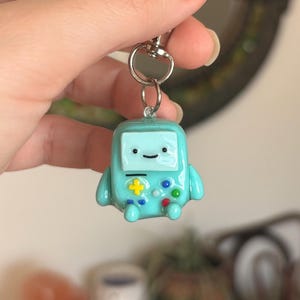 May include: A teal-colored keychain featuring a cartoon character with a square head and a smiling face. The character has a yellow plus sign and colorful buttons, resembling a video game controller. The keychain is attached to a silver clasp.