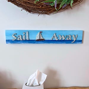 May include: A rectangular wooden sign with the words "Sail Away" in silver lettering, and a sailboat in the center. The background is painted blue, with a brown wreath and lavender sprigs in the background.