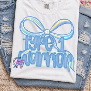May include: A white short-sleeved t-shirt featuring a large blue and yellow ribbon graphic with the text type 1 warrior in blue bubble letters. The shirt is layered over a pair of distressed blue denim jeans with frayed holes and a visible tag.