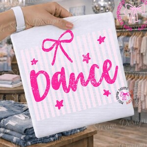 May include: White t-shirt with a pink bow, vertical stripes, and the word "Dance" in a distressed font. Pink stars are scattered around the text. The shirt is held up by a person.
