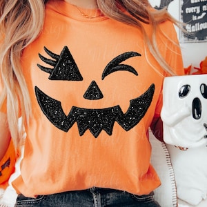 May include: An orange t-shirt featuring a large, black, glittery jack-o'-lantern face graphic with a winking eye and a jagged smile. The shirt is paired with dark gray denim jeans and a white ceramic mug shaped like a ghost with a small mouse.