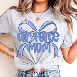 May include: A light gray t-shirt with the words "Air Force Mom" in blue script, accented by a blue bow design. The shirt is paired with denim shorts.