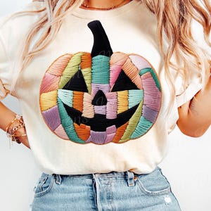 May include: Cream-colored t-shirt featuring a colorful, embroidered pumpkin design. The pumpkin has a black stem and a jack-o'-lantern face with triangular eyes and a smiling mouth. The pumpkin is made of various colors, including orange, green, purple, and yellow.