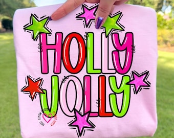 May include: A light pink sweatshirt with the words "HOLLY JOLLY" in a colorful, festive design. The text is outlined in black and filled with red, green, and pink. Star graphics in various colors surround the text, creating a cheerful holiday theme.