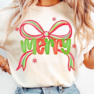Könnte beinhalten: Cremefarbenes T-Shirt mit einem großen, festlichen Schleifendesign in Rot und Grün, mit dem Wort "MERRY" in grünen und roten Buchstaben. Kleine Schneeflocken-Akzente umrunden die Schleife. Das Shirt wird mit hellblauen Jeans kombiniert.