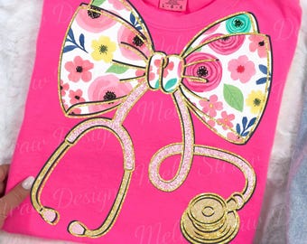 Pode incluir: Camiseta rosa vibrante com um design de laço floral e estetoscópio. O laço apresenta flores rosa, brancas e amarelas. O estetoscópio é dourado com detalhes brilhantes. Marca Comfort Colors.
