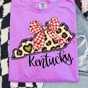 Peut inclure: T-shirt lilas Comfort Colors avec un contour de l'État du Kentucky, un imprimé léopard et un nœud vichy rouge et blanc. Le mot "Kentucky" est écrit en cursive sous le contour de l'État.