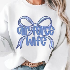 May include: A white sweatshirt with a blue bow design and the words "Air Force Wife" in a matching blue font. The design is centered on the front of the long-sleeved top. The sweatshirt is made of a soft material.