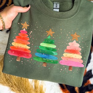 Puede incluir: Una sudadera verde oliva con tres árboles de Navidad coloridos, cada uno coronado con una estrella dorada con purpurina. Los árboles tienen un patrón de puntos de purpurina rosa claro y dorado.