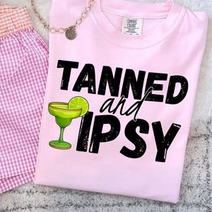 May include: A light pink t-shirt with the words "TANNED and TIPSY" in black lettering, along with a margarita graphic. The shirt is paired with pink gingham shorts and a silver necklace.
