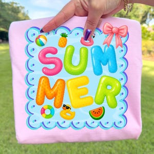 May include: A light pink t-shirt featuring the word "SUMMER" in vibrant bubble letters. The design includes a beach ball, pineapple, inflatable ring, sunglasses, watermelon slice, and a pink bow. The shirt has a scalloped border.