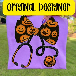 May include: A lilac t-shirt with a black bow decorated with orange jack-o'-lanterns and a stethoscope design. The text "ORIGINAL DESIGNER" is in a yellow rectangle. The bow and stethoscope have glitter detailing.