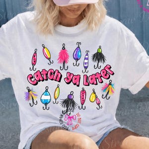 May include: White t-shirt with a colorful graphic of fishing lures and the text "Catch ya Later" in pink. The lures are in various colors, including pink, yellow, blue, and black. A pink bow is also visible.
