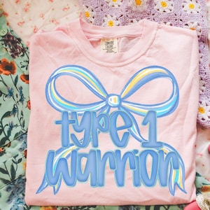 May include: A light pink t-shirt with a blue and yellow bow design and the words "Type 1 Warrior" in blue. The shirt is made of a soft material and is suitable for casual wear.