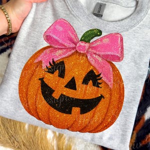 May include: A light gray sweatshirt featuring a glittery orange pumpkin with a pink bow and eyelashes. The pumpkin has a smiling face and a green stem. The sweatshirt is a crew neck style.