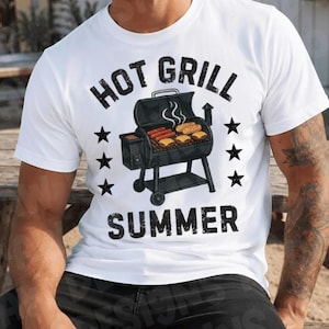 May include: A white short-sleeved t-shirt featuring a black graphic of a charcoal grill with food cooking on the grate. The text "HOT GRILL SUMMER" is printed in black above and below the grill illustration, flanked by black stars.