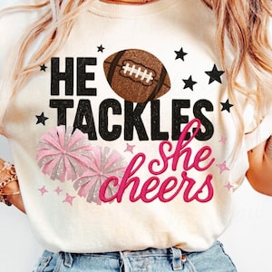 Può includere: T-shirt color crema con la scritta "He Tackles, She Cheers" in nero e rosa. Il design include un pallone da football, stelle e pompon rosa, perfetto per i fan del football.