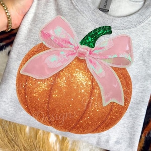 May include: A light gray sweatshirt featuring a large, glittery orange pumpkin with a pink bow and green stem. The pumpkin design is the focal point, perfect for fall or Halloween. The sweatshirt is a crew neck style.