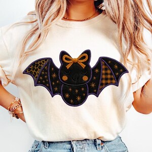 Puede incluir: Una camiseta color crema con un diseño de murciélago con temática de Halloween. El murciélago tiene un cuerpo negro con estrellas doradas, un lazo dorado y alas estampadas con estampado de leopardo y cuadros. El diseño está delineado en morado.