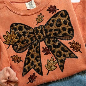 Puede incluir: Una sudadera naranja con un gran lazo con estampado de leopardo y hojas de otoño bordadas. El lazo es el punto focal, con costuras detalladas y un patrón de leopardo clásico. Las hojas son en tonos dorados, marrones y rojos.