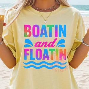 May include: A light yellow t-shirt with the words "BOATIN and FLOATIN" in colorful, tie-dye style lettering. The shirt features a wavy blue line design at the bottom. The shirt is a casual, beach-themed apparel item.