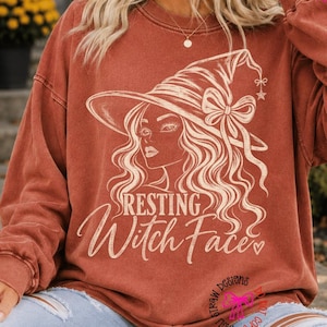 May include: A rust-coloured long-sleeve crewneck sweatshirt featuring a white line-art graphic of a person wearing a witch hat with a bow. The text "RESTING Witch Face" is printed below the graphic in a stylised script and serif font.