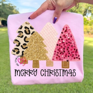 Op de afbeelding: Een lichtroze t-shirt met drie kerstboomontwerpen. De bomen hebben luipaardprint, gouden glitter, roze en wit patroon en pailletten texturen. De woorden "MERRY CHRISTMAS" staan in het zwart onderaan.