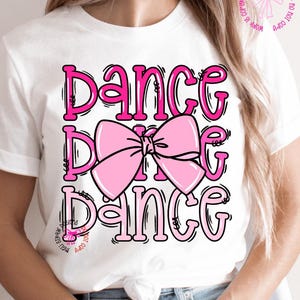 May include: White t-shirt with the word "DANCE" in pink, repeated three times, with a pink bow graphic in the center. The text has a black outline. The shirt is a casual style.