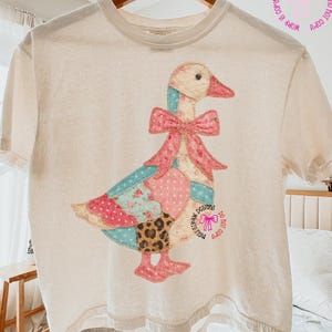 May include: A cream-coloured t-shirt with a patchwork goose design. The goose has a pink bow and a body made of various patterned fabrics in pink, blue, and leopard print. The text "MEW DESIGNS" is visible on the shirt.