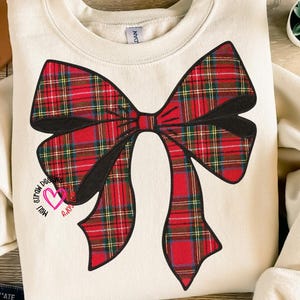 May include: Cream-colored sweatshirt featuring a large, red plaid bow design with black outlines. The bow has green and yellow accents. The text "Meli's Straw Designs" and a pink heart are printed on the lower left side of the bow.