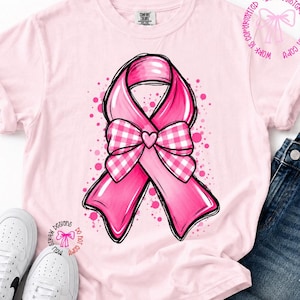 Op de afbeelding: Een lichtroze T-shirt met korte mouwen en een grote afbeelding van een roze bewustzijnslint met een wit-roze geruite strik in het midden. Het ontwerp bevat verspreide roze stippen en een klein cirkelvormig logo in de rechterbovenhoek.