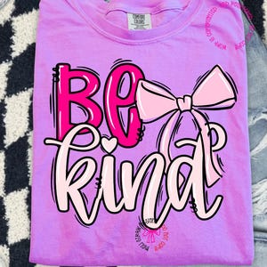 May include: A lilac Comfort Colors t-shirt with the words "BE KIND" in a bubble font, accented with a pink bow graphic. The text and bow are outlined in black. The shirt is folded.